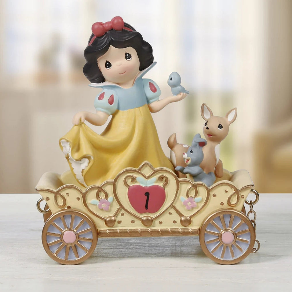 A decorative figurine of Snow White in a yellow dress seated in a gold-toned carriage with pink accents, accompanied by a small bird, rabbit, and a tan animal, photographed against a softly blurred indoor background.