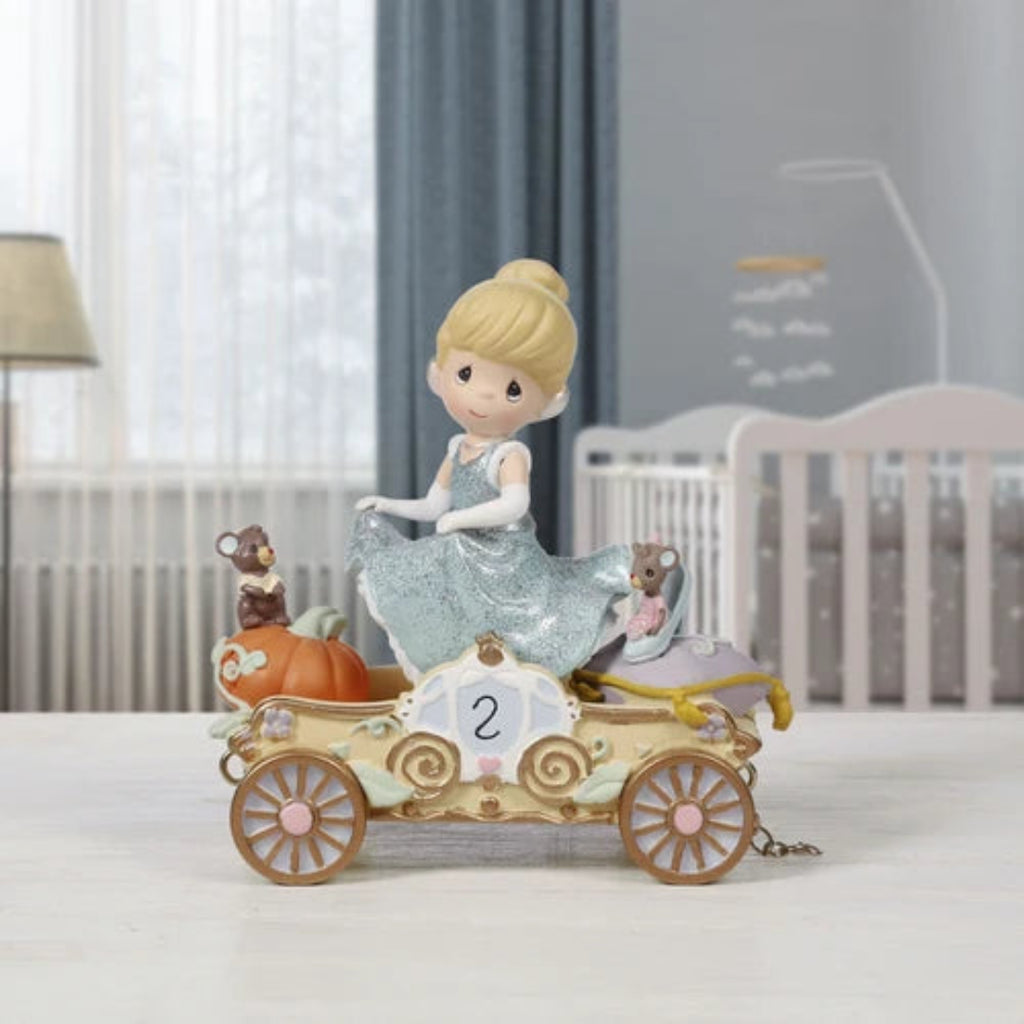 A Cinderella-themed figurine sits on a white tabletop in a softly blurred nursery setting. The decorative car features a blonde Cinderella figure in a shimmering light blue gown, with small mice, a pumpkin, and pastel floral details on the carriage.