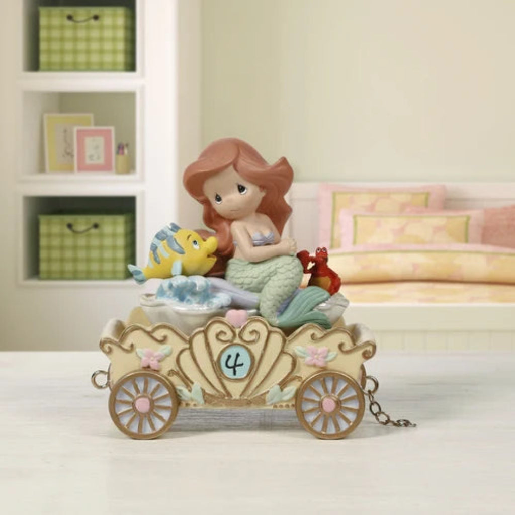 A mermaid-themed carriage figurine featuring a mermaid with reddish-brown hair, a green tail, a yellow fish, and a red crab sitting in an ornate cream-colored carriage with pink accents, displayed on a tabletop with a softly blurred bedroom background.