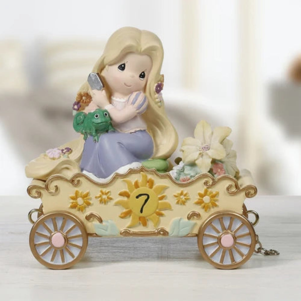 A Precious Moments figurine of Rapunzel sitting in her golden carriage, holding a green frog and looking out of the window. The carriage is cream-colored with gold accents, a yellow sun emblem with the number 7, and decorated with flowers.