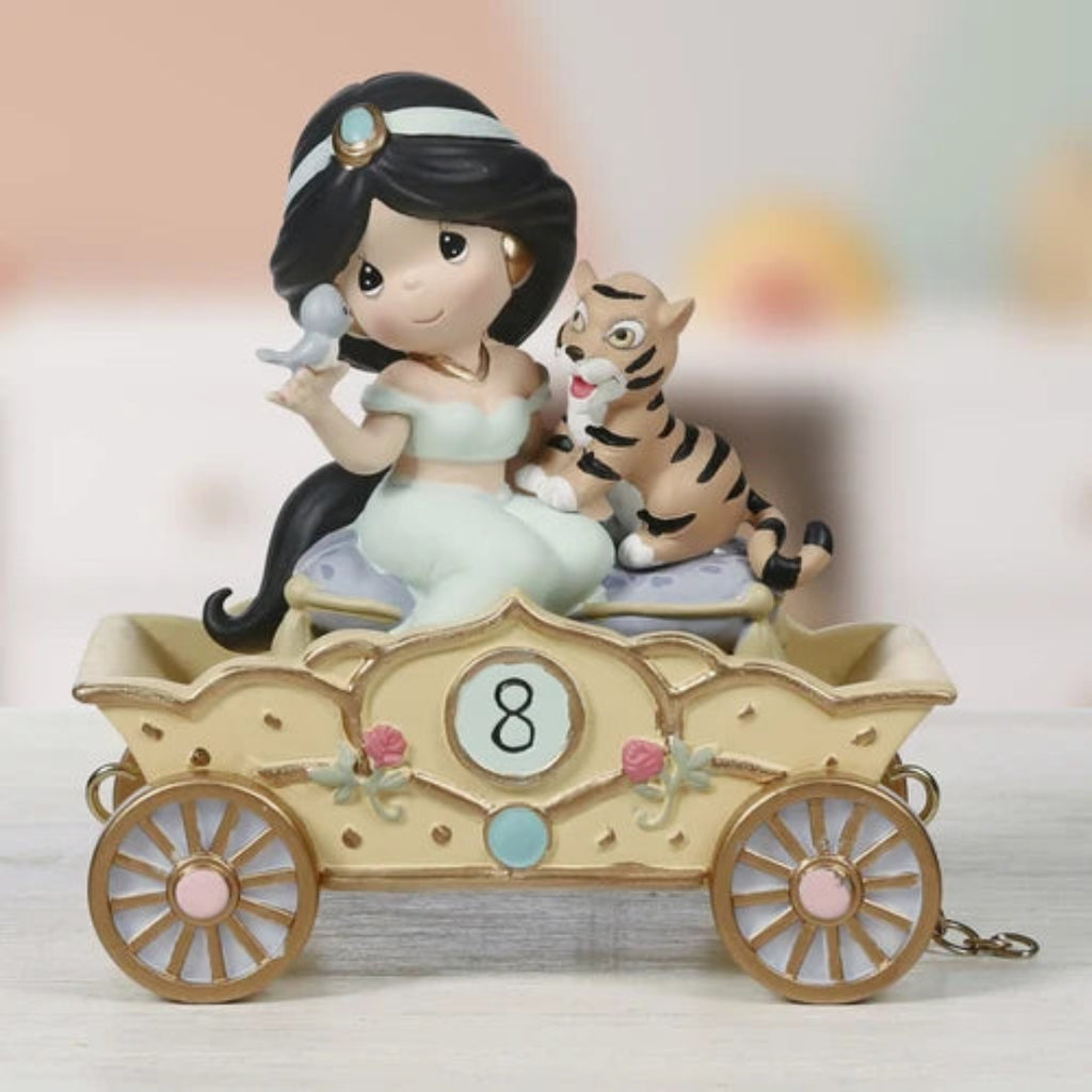 A decorative figurine of a woman in a flowing white dress seated in a beige carriage with gold wheels, accompanied by a small tiger. The figurine is photographed on a light surface against a softly blurred pastel background.