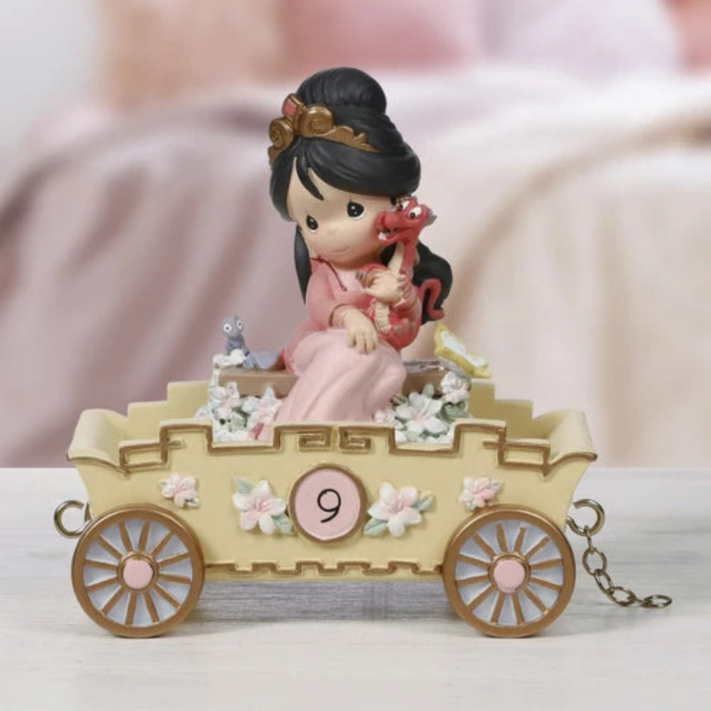 A Precious Moments figurine of a woman in a pink robe sitting in a beige carriage decorated with flowers, holding a red dragon, displayed on a light surface with a soft pink blurred background.