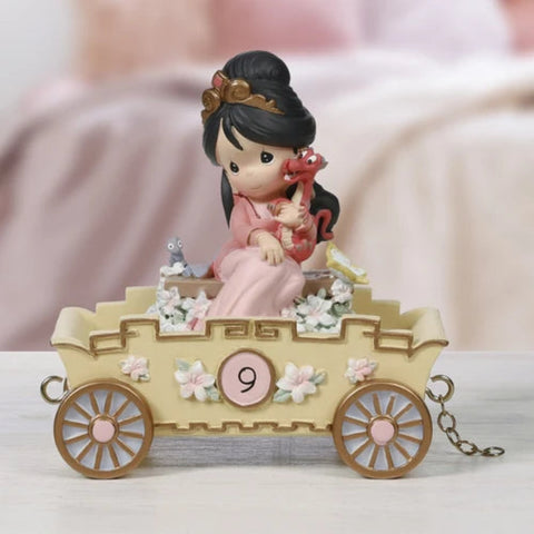 A Precious Moments figurine of a woman in a pink robe sitting in a beige carriage decorated with flowers, holding a red dragon, displayed on a light surface with a soft pink blurred background.