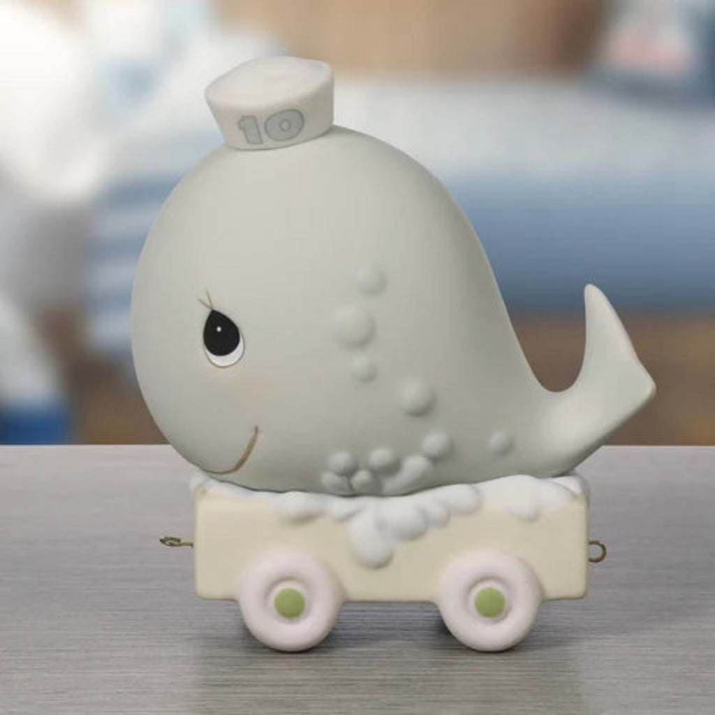 A small whale-shaped decorative object on a cream-colored cart with white wheels, photographed on a gray tabletop with a softly blurred indoor background.