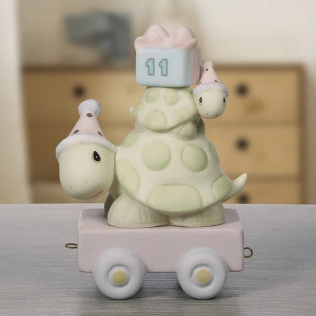 A ceramic turtle figurine sitting on a small pink cart, with a pastel gift box on its shell and party hats on its head and tail. The figurine is photographed on a tabletop with a softly blurred indoor background.