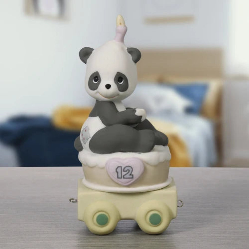 A cute panda figurine sitting on a decorated cake-shaped base with a pink heart that says '12' and a small horn on top, displayed on a tabletop with a blurred bedroom background.