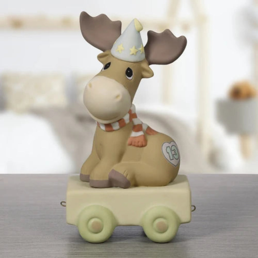 A ceramic moose figurine sitting on a small cream-colored toy cart, wearing a white party hat with yellow stars and an orange-and-white striped scarf. The moose is tan with brown antlers and hooves, and a white heart emblem is printed on its side.