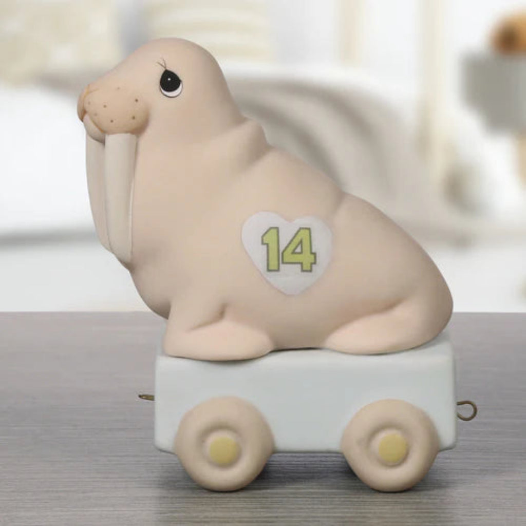 A walrus figurine sitting on a small white cart with wheels, photographed on a gray tabletop with a softly blurred indoor background. The walrus is light beige with a white heart-shaped patch on its side showing the number 14 in yellow-green.