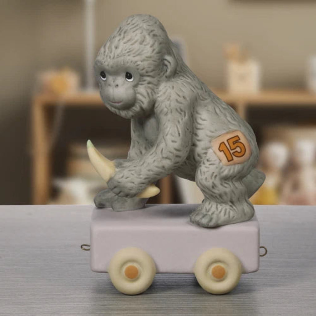 A small gray gorilla figurine holding a yellow trident, standing on a pale lavender toy cart with cream wheels. The side of the cart has a peach patch with the number 15.
