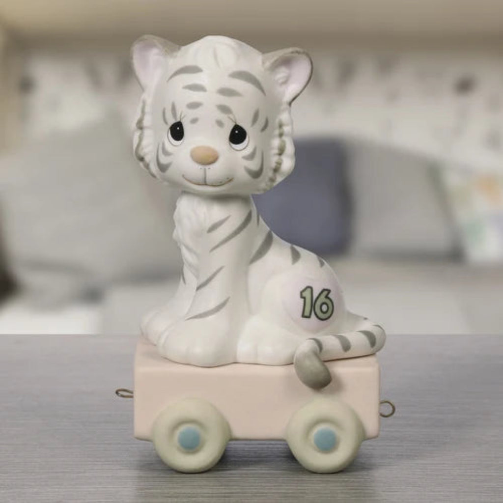A white tiger figurine with gray stripes sitting on a small pale pink toy car with wheels, photographed on a tabletop with a softly blurred indoor background.