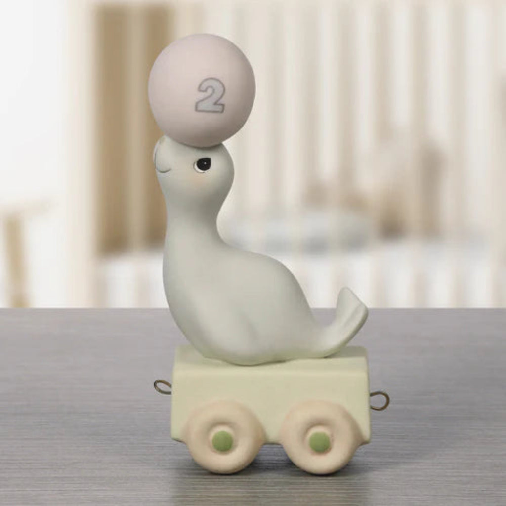 A small decorative figurine of a white seal sitting on a cream-colored toy cart with two wheels, balancing a round beige ball marked with the number 2 on its forehead. The figurine is placed on a gray wooden surface with a softly blurred indoor background.