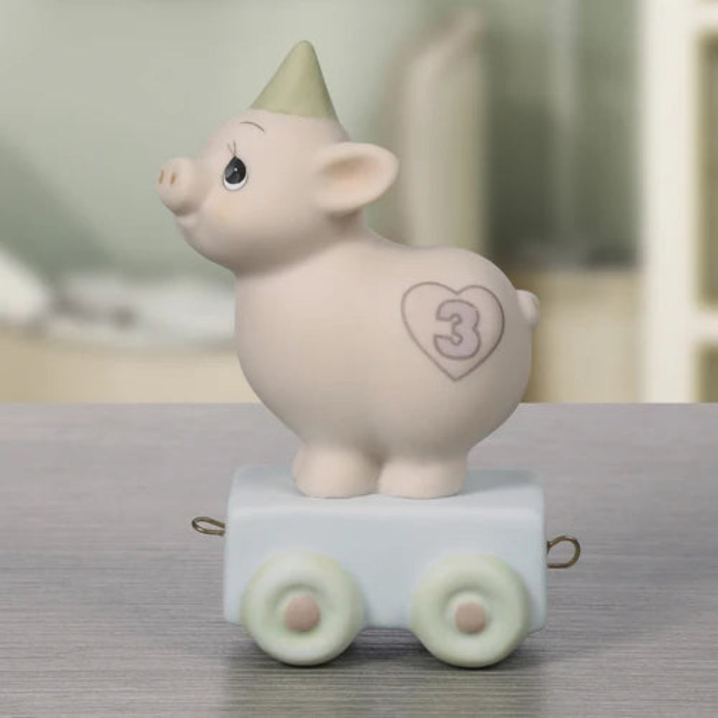 A ceramic pig figurine wearing a pointed party hat, standing on a small white cart with pale green wheels. The pig has a heart-shaped outline with the number 3 painted on its back.