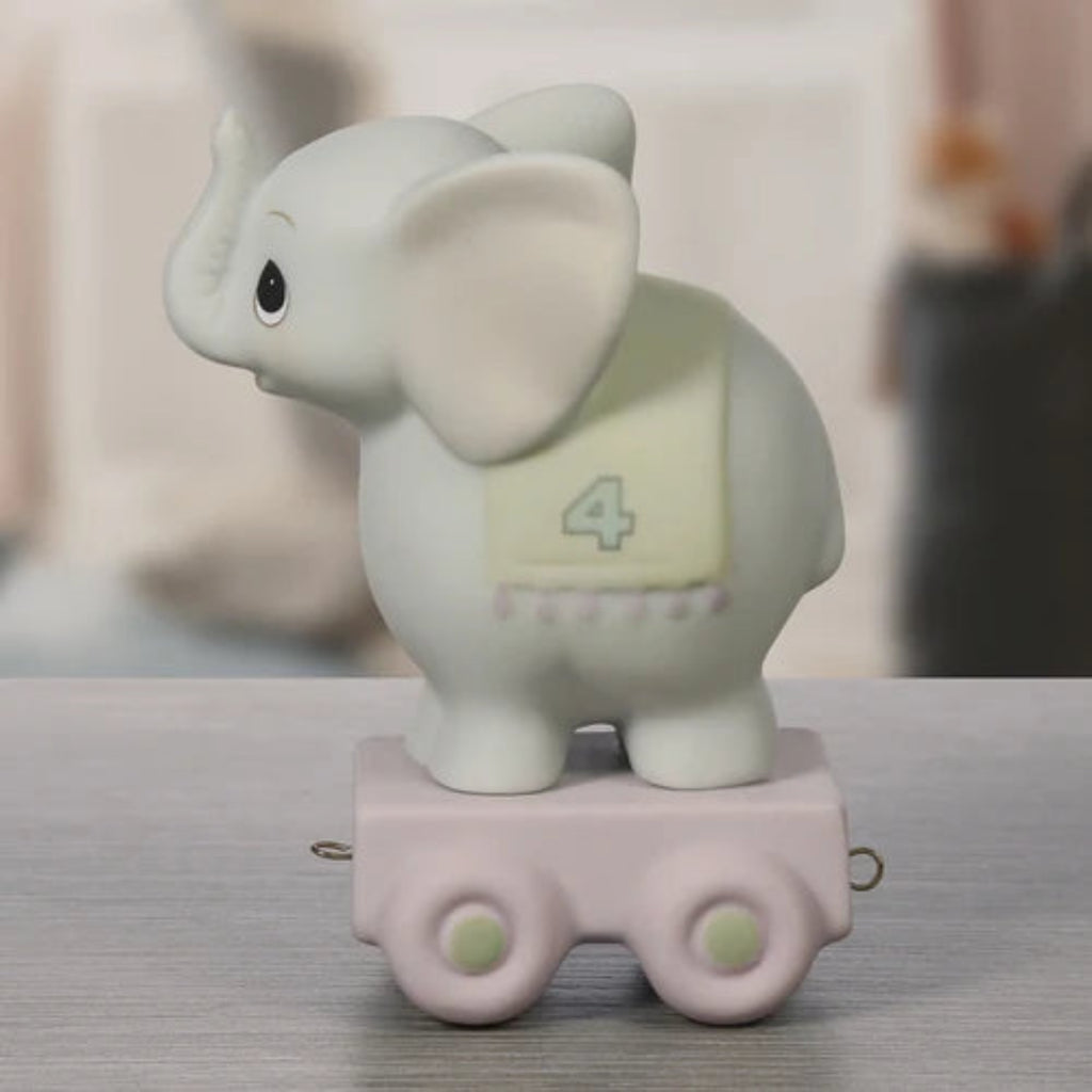 A small elephant figurine standing on a pink toy cart, photographed on a tabletop with a softly blurred indoor background. The elephant is off-white with a pale yellow patch showing the number 4, and the cart is light pink with pale green wheel details.