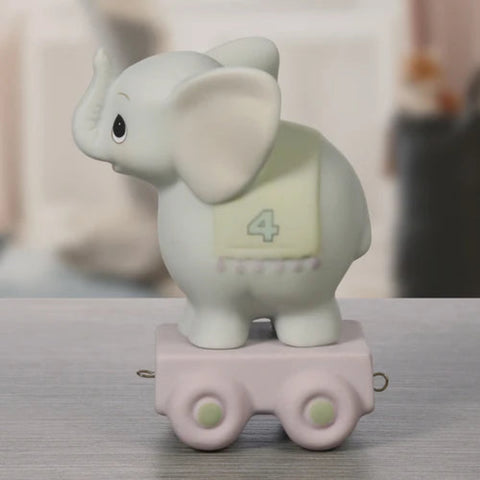 A small elephant figurine standing on a pink toy cart, photographed on a tabletop with a softly blurred indoor background. The elephant is off-white with a pale yellow patch showing the number 4, and the cart is light pink with pale green wheel details.