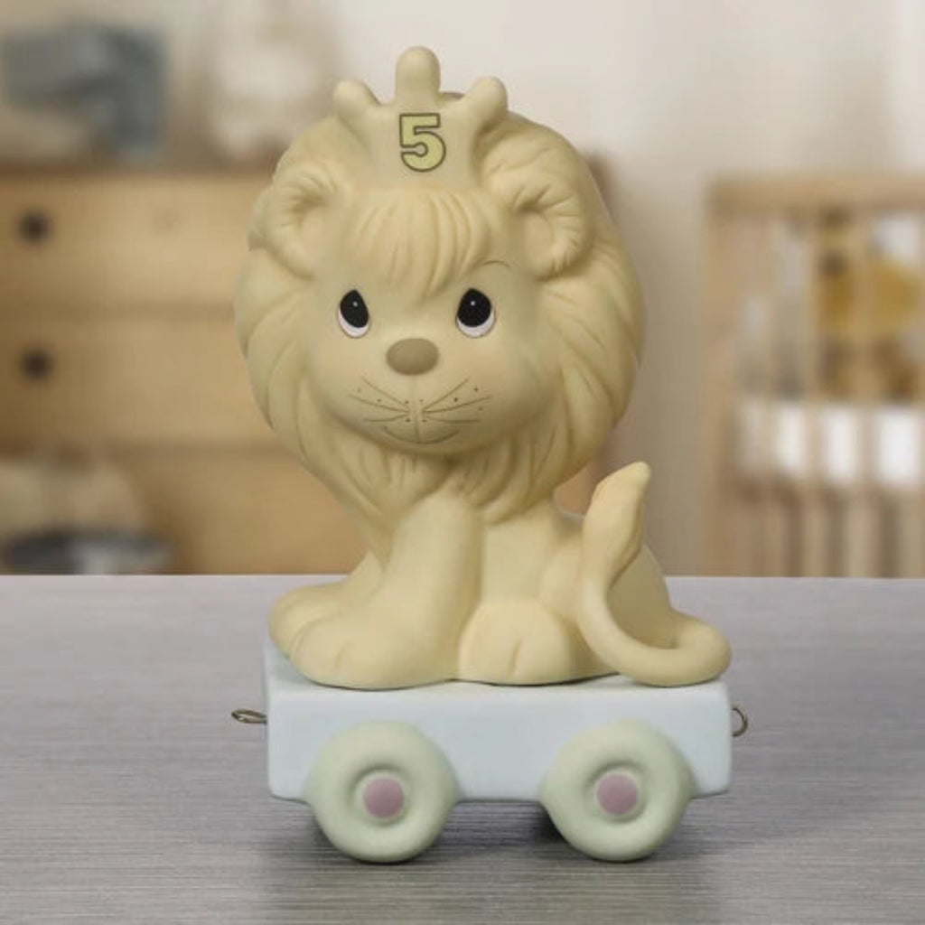 A cute cream-colored lion figurine sitting on a small white toy cart with pastel green wheels, featuring a gold '5' on its forehead, displayed on a gray tabletop with a blurred nursery background.