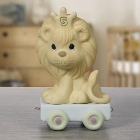 A cute cream-colored lion figurine sitting on a small white toy cart with pastel green wheels, featuring a gold '5' on its forehead, displayed on a gray tabletop with a blurred nursery background.