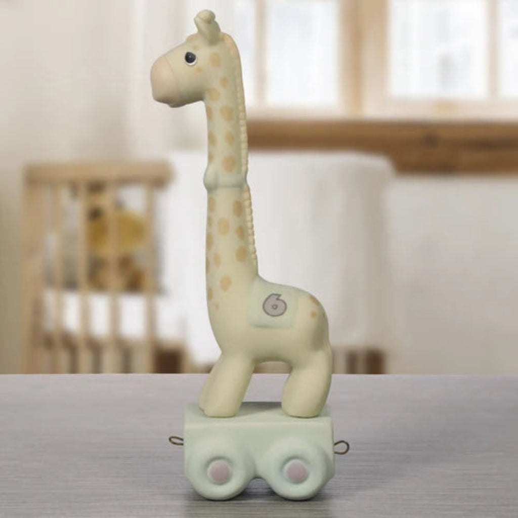 A ceramic giraffe toy on a small wheeled base, standing on a gray tabletop with a softly blurred nursery background.