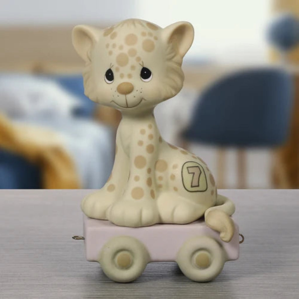 A ceramic figurine of a cute cream-colored spotted kitten sitting on a small pastel pink toy cart, displayed on a gray tabletop with a blurred indoor background.