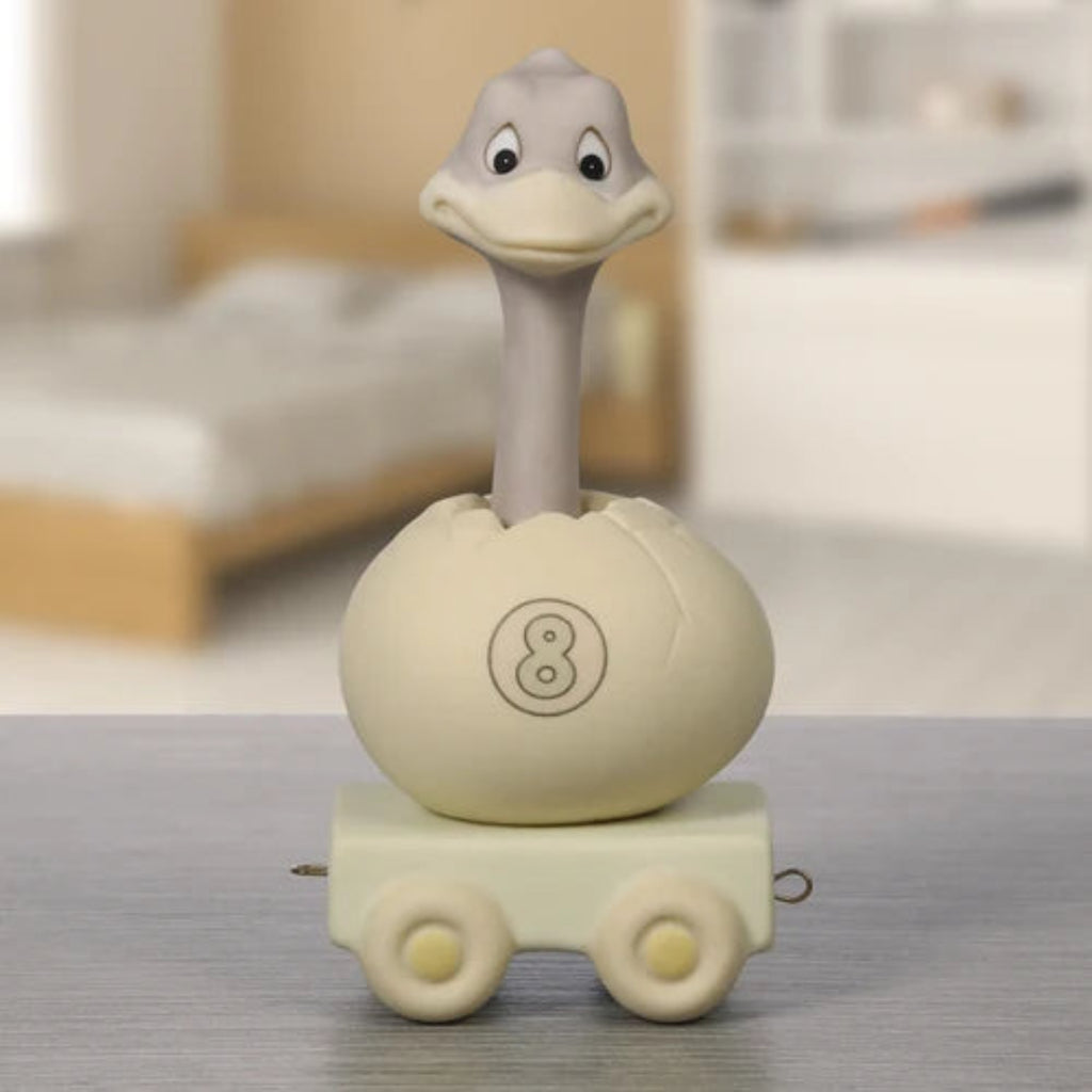A novelty toy or novelty container shaped like a duckling emerging from an egg on a small wheeled base, photographed standing on a tabletop with a softly blurred bedroom background.