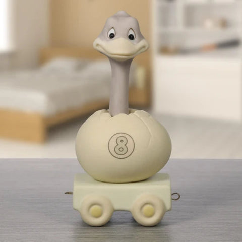 A novelty toy or novelty container shaped like a duckling emerging from an egg on a small wheeled base, photographed standing on a tabletop with a softly blurred bedroom background.
