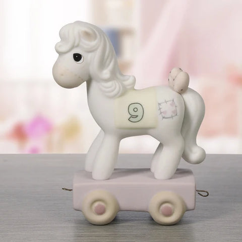 A white ceramic rocking horse toy on a pastel pink and lavender wheeled base, photographed on a gray tabletop with a softly blurred pastel background.