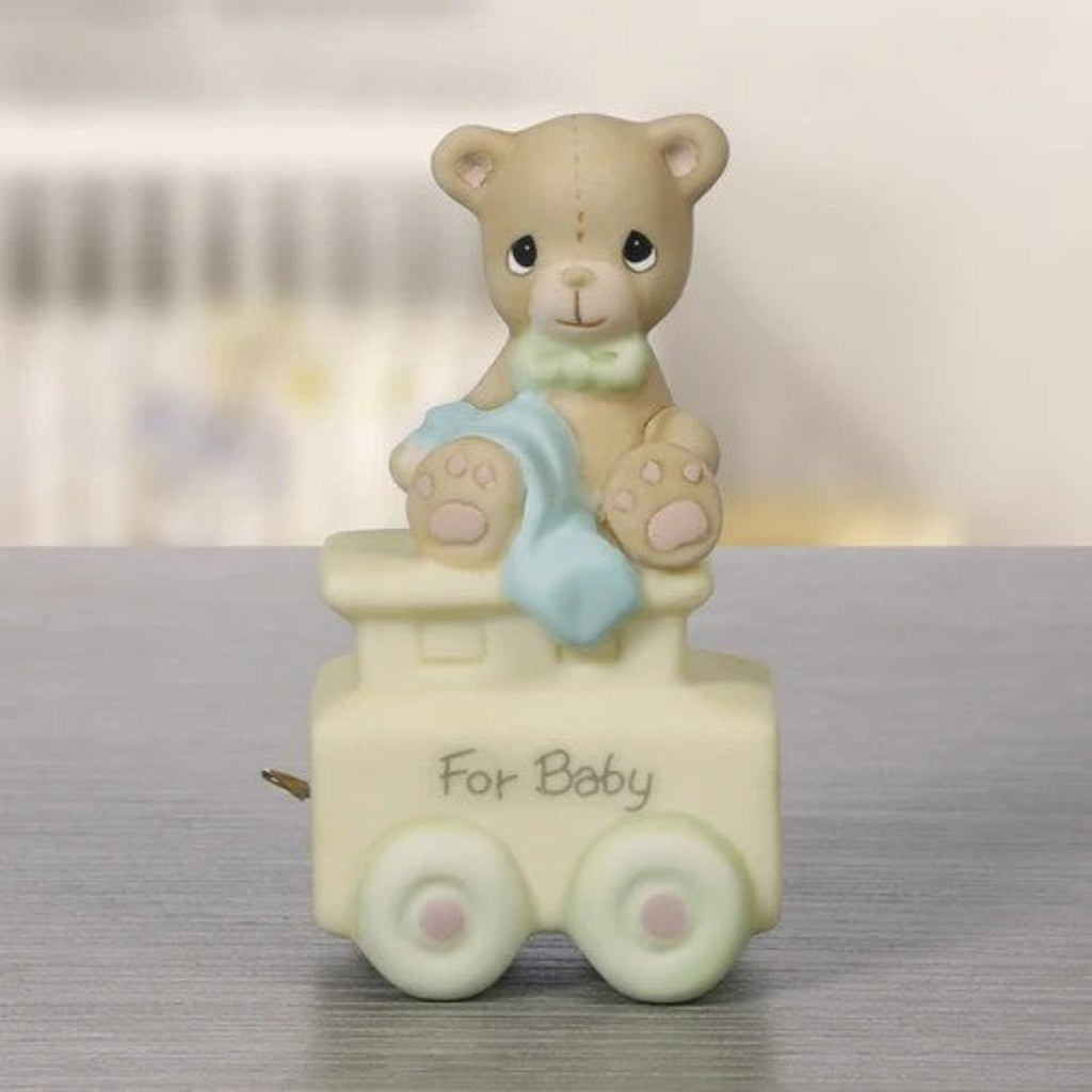 A ceramic teddy bear figurine sitting on a small train car with the words 'For Baby' written on the side. The bear is beige with a light blue blanket and a green bow tie, while the train car is cream with pale green wheels.