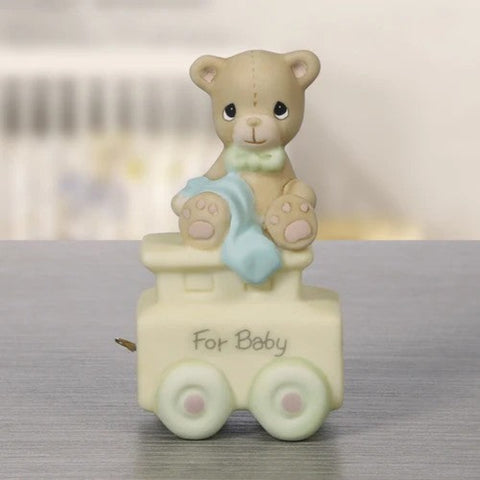 A ceramic teddy bear figurine sitting on a small train car with the words 'For Baby' written on the side. The bear is beige with a light blue blanket and a green bow tie, while the train car is cream with pale green wheels.