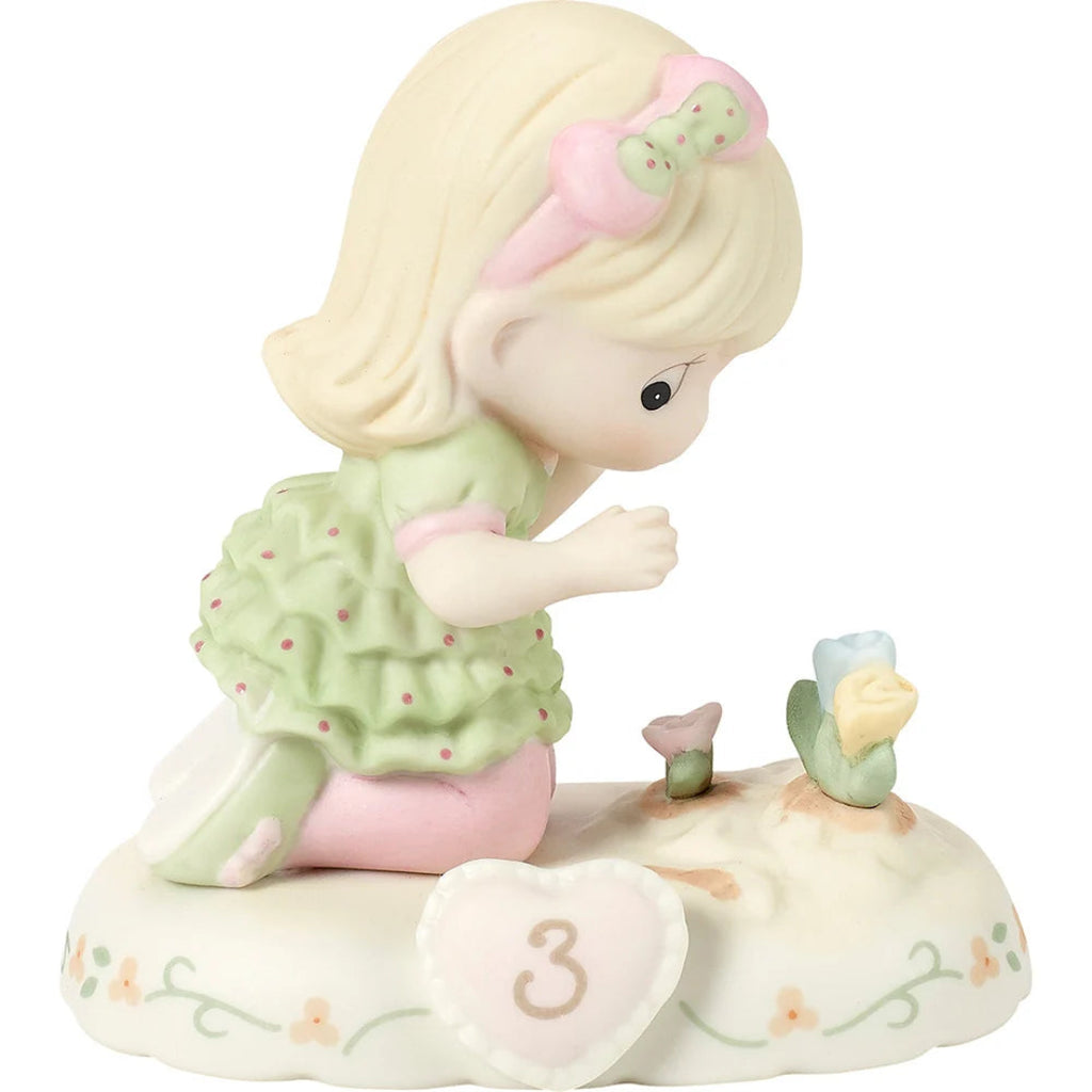 A porcelain figurine of a blonde girl kneeling beside small flowers on a decorative base with a heart-shaped ornament marked '3', shown on a plain white background.