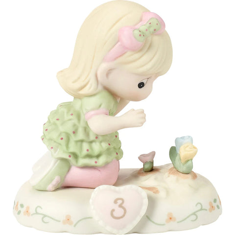 A porcelain figurine of a blonde girl kneeling beside small flowers on a decorative base with a heart-shaped ornament marked '3', shown on a plain white background.