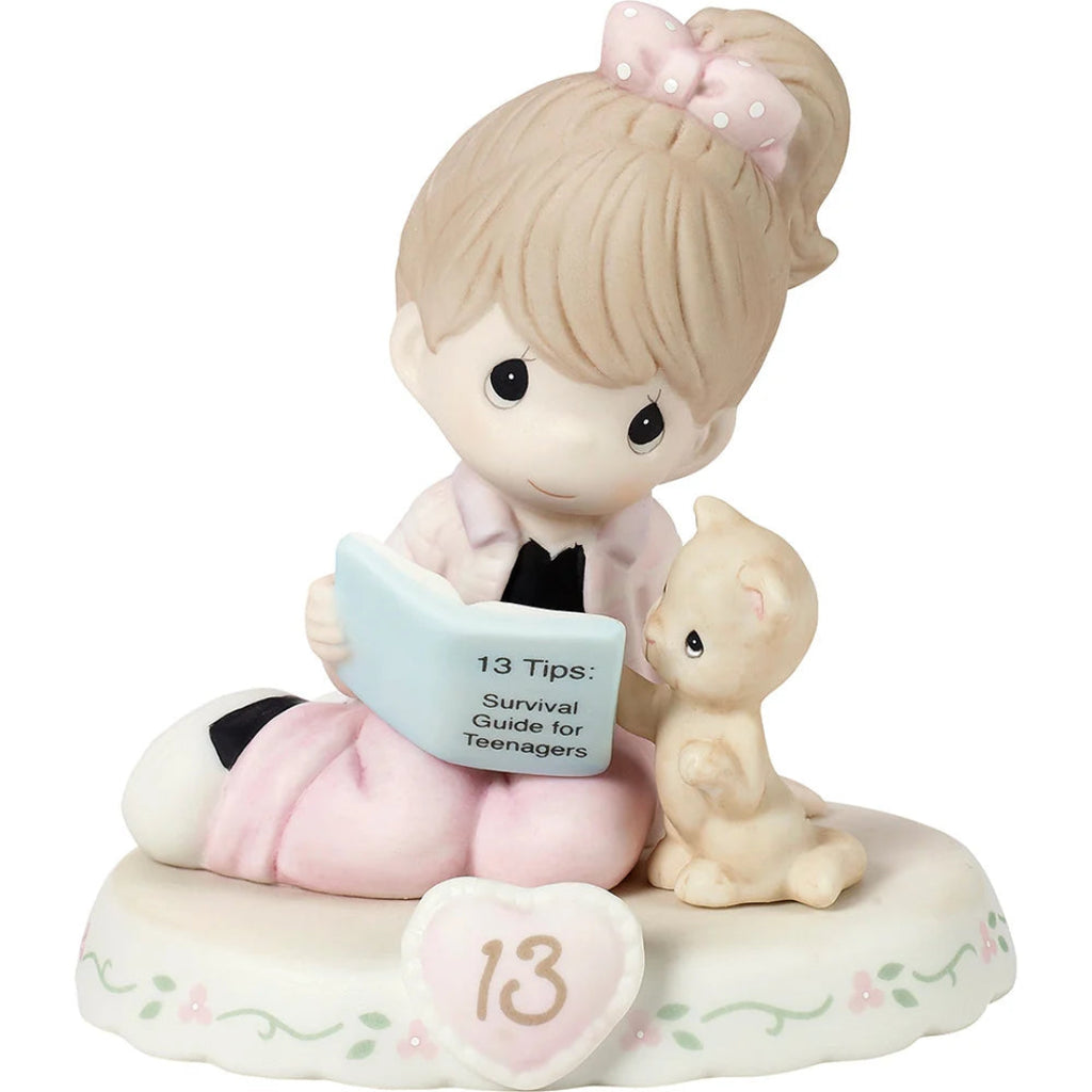 A Precious Moments figurine of a girl with brunette hair in a pink bow, sitting and reading a book next to a small cat, on a white floral base with a heart-shaped ornament with 13 on it