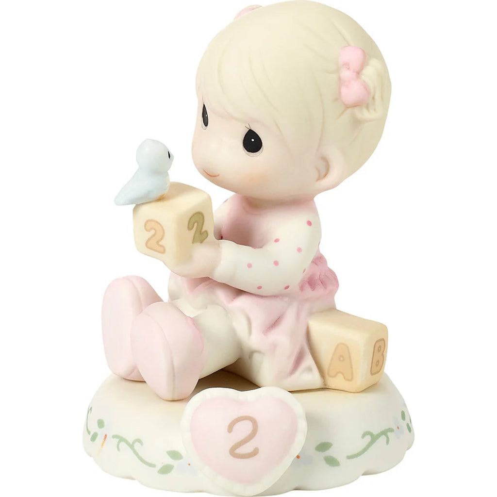 A Precious Moments figurine of a baby girl with blonde hair and a pink bow, sitting on a base with a heart-shaped number 2, holding blocks with the number 2 and letters A and B, with a small white bird on her block.