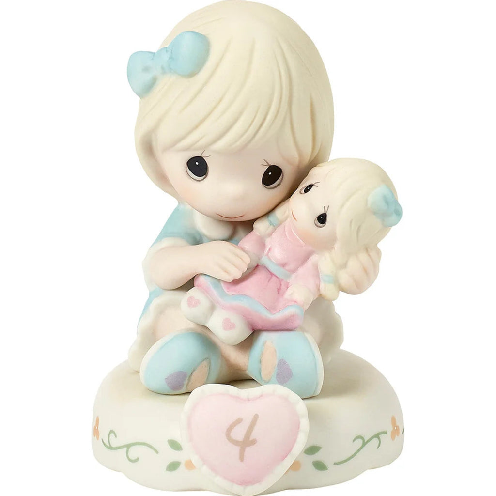 A Precious Moments figurine of a blonde girl hugging a smaller doll, both with light blue bows, sitting on a cream-colored base with a pink heart-shaped ornament marked with the number 4.