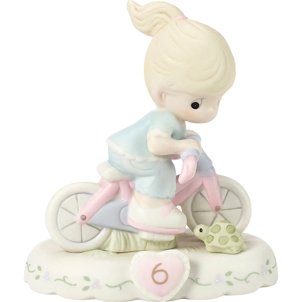 A ceramic figurine of a young girl with blonde hair in a ponytail riding a pink and white bicycle on a decorative base. The figurine features a small green turtle and a pink heart-shaped ornament with the number 6.