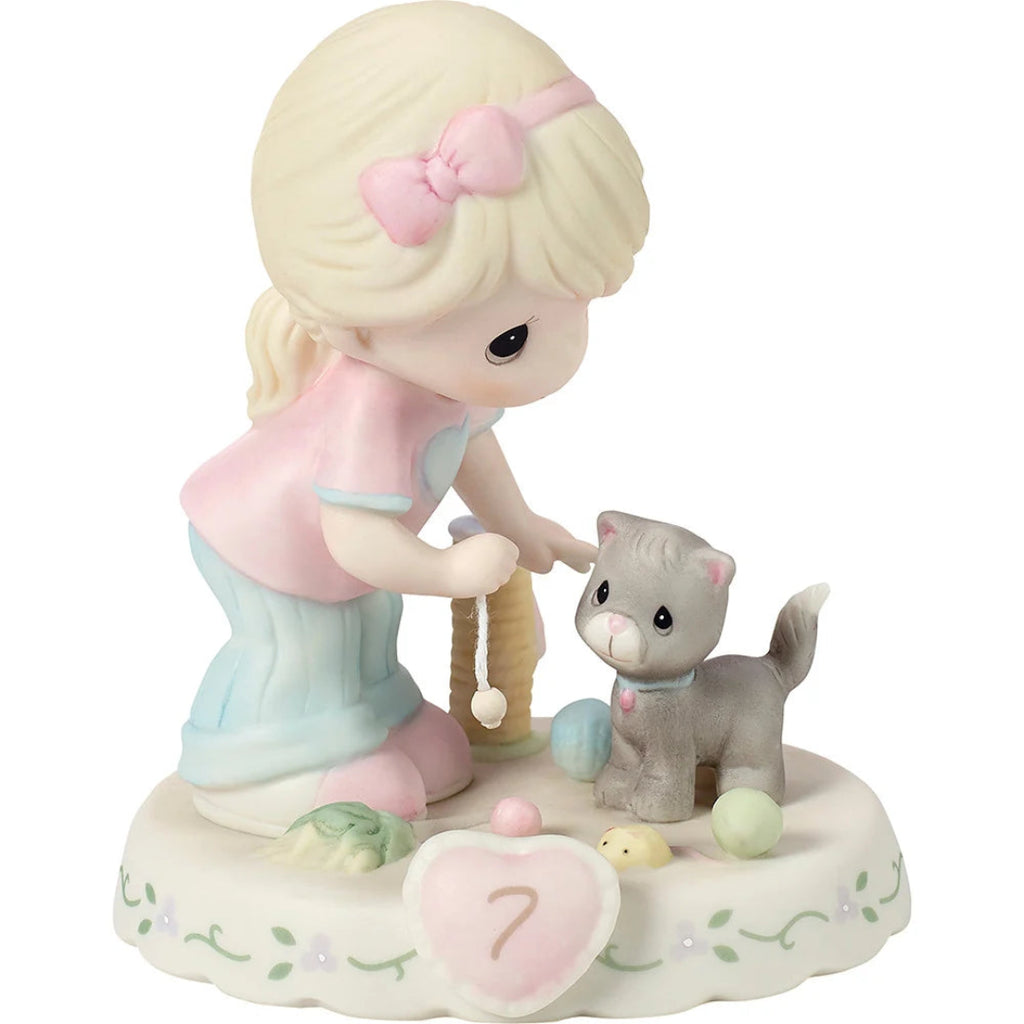 A Precious Moments figurine featuring a blonde girl with a pink bow kneeling beside a gray kitten, with small pastel toy balls and a heart-shaped plaque on a decorative base.