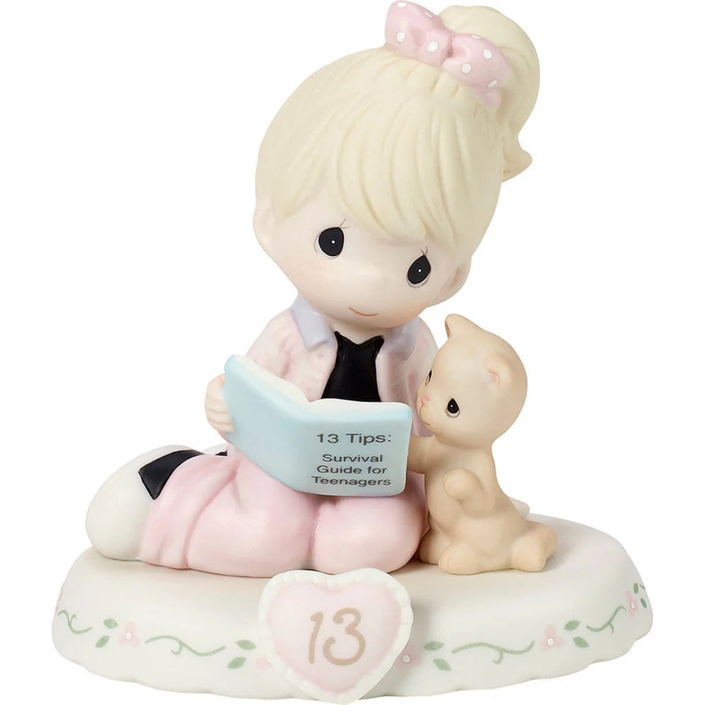 A Precious Moments figurine of a girl with blonde hair in a pink bow, sitting and reading a book next to a small cat, on a white floral base with a heart-shaped ornament showing the number 13.