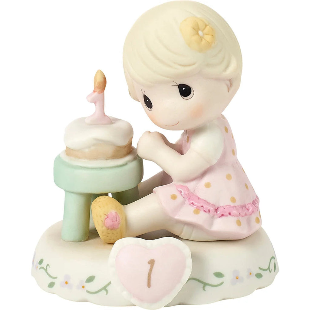 A ceramic figurine of a young girl with blonde hair and a yellow flower in her hair, sitting beside a small cake with a single candle on a white base. She wears a pink polka-dot dress, and a pink heart-shaped plaque with the number one is in front of her.