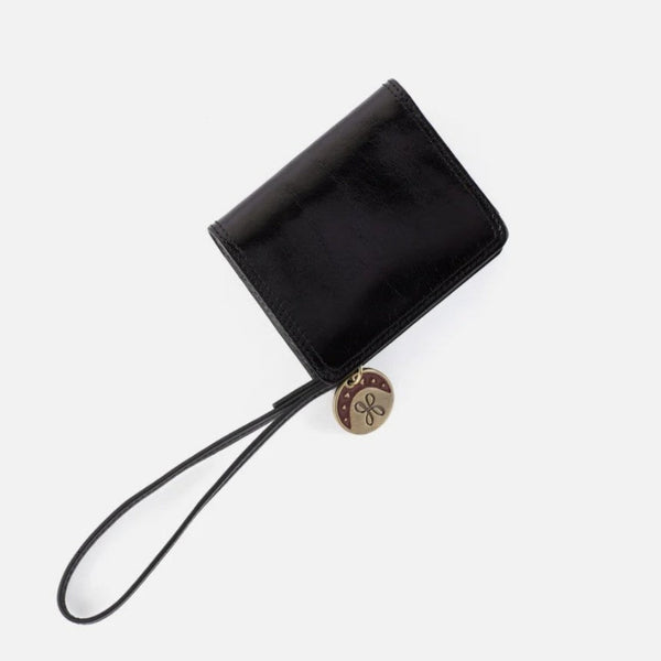 A black leather wristlet wallet with a matching wrist strap and a round gold-tone charm featuring a stylized 'S' logo, shown on a light gray background.