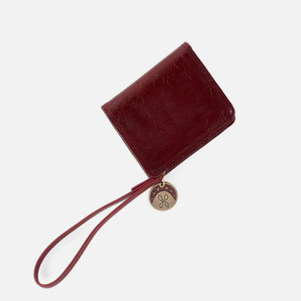 A dark red leather wristlet wallet with a matching wrist strap and a round gold-tone metal charm, shown on a light gray background.