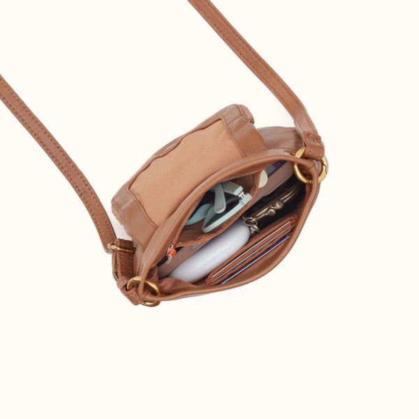 A tan leather crossbody bag shown open, revealing its interior compartments filled with cards, sunglasses, and a wallet, on a solid off-white background.