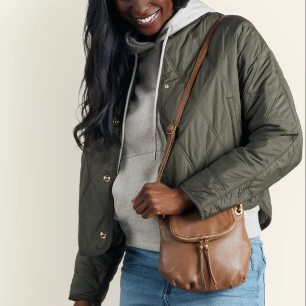 A woman wearing an olive green quilted puffer jacket over a gray hoodie and blue jeans, carrying a brown leather crossbody bag with a gold-tone clasp and tassel detail.