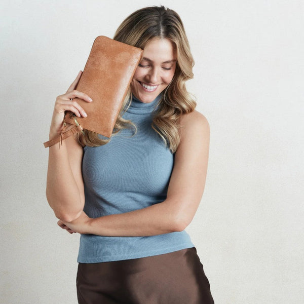 A woman holding a tan leather clutch purse against her face, wearing a light blue ribbed sleeveless top and brown satin pants, set against a plain light gray background.