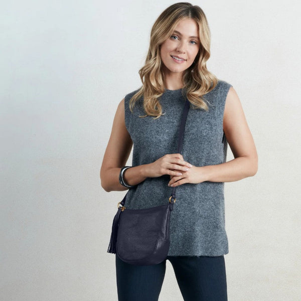 A woman wearing a sleeveless heather gray knit top and dark pants, carrying a dark navy blue leather crossbody bag with gold-tone hardware against a light gray studio background.