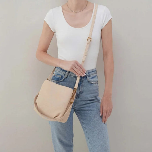A woman wearing a white scoop-neck t-shirt and light blue jeans carries a beige leather shoulder bag with gold-tone hardware against a plain light gray background.