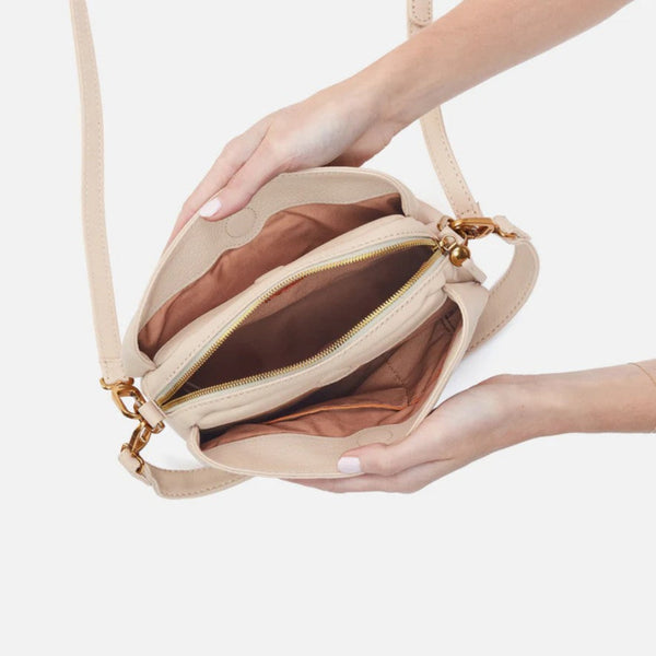 A beige crossbody handbag with gold-tone hardware, held open by two hands to show the interior space and brown lining against a light gray background.