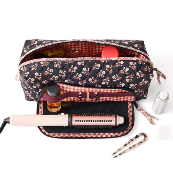 A quilted floral makeup bag shown open on a white background, with a curling iron, tweezers, and other small accessories placed inside and around it.