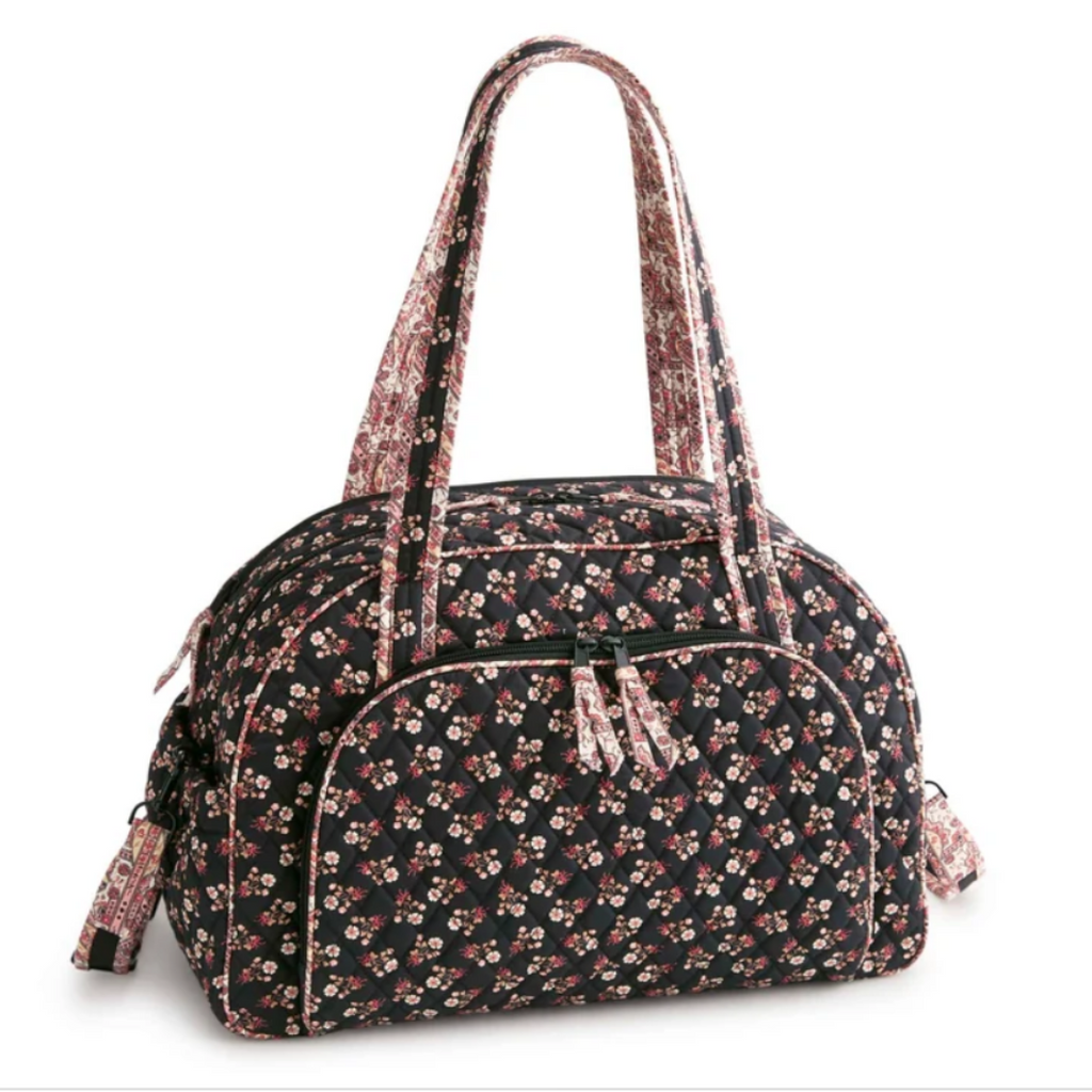 A floral print duffel bag with a black background, featuring small pink, red, and white flowers. It has dual top handles, a front zippered pocket, and a matching shoulder strap, shown on a plain white background.