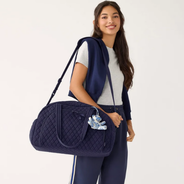A woman wearing a gray t-shirt, navy cardigan, and navy pants carries a navy blue quilted duffel bag with a blue and white patterned keychain charm against a plain white studio background.