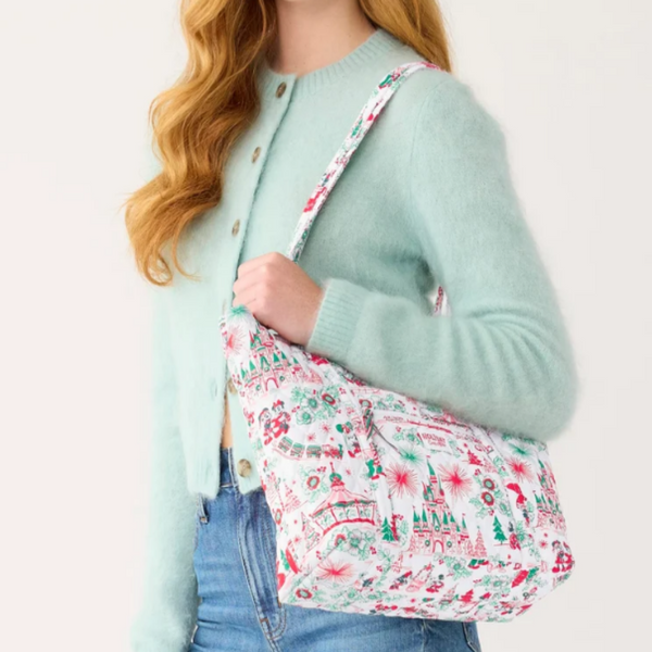 A woman wearing a mint green cardigan and blue jeans carries a white shoulder bag with a colorful Christmas village pattern featuring red, green, and blue details.