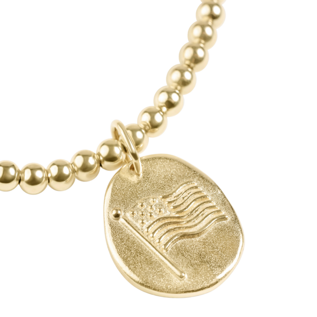 A gold beaded bracelet featuring a textured, irregularly shaped round pendant engraved with an American flag design, shown against a solid white background.
