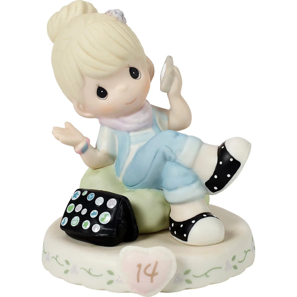 A ceramic figurine of a blonde girl sitting on a green stool and holding a phone, with a black polka-dot purse beside her. The figurine is mounted on a round white floral base decorated with a pink heart and the number 14.